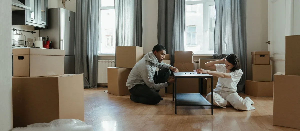 Zwei Personen bauen in einer Wohnung voller Umzugskartons einen kleinen Tisch zusammen
