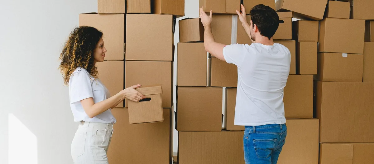 Zwei Personen stapeln unbedruckte Umzugskartons in einem leergeräumten Zimmer an einer weißen Wand