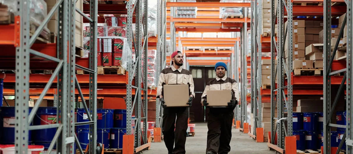 Zwei Mitarbeiter in Arbeitskleidung tragen gemeinsam einen Karton durch einen Lagergang zwischen hohen Regalen
