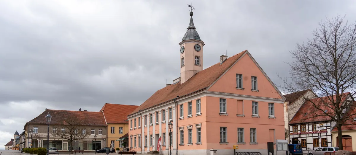 Marktplatz einer kleinen Landstadt mit rosafarbenem Rathaus, Uhrturm und niedrigen Häusern mit roten Ziegeldächern