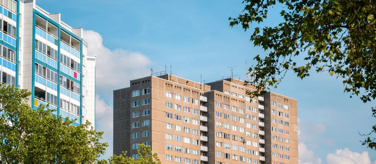 Zwei Wohnhochhäuser in Plattenbauweise, eines blau verkleidet, das andere mit beigen Fassadenplatten und Loggien, dahinter Laubbäume und blauer Himmel