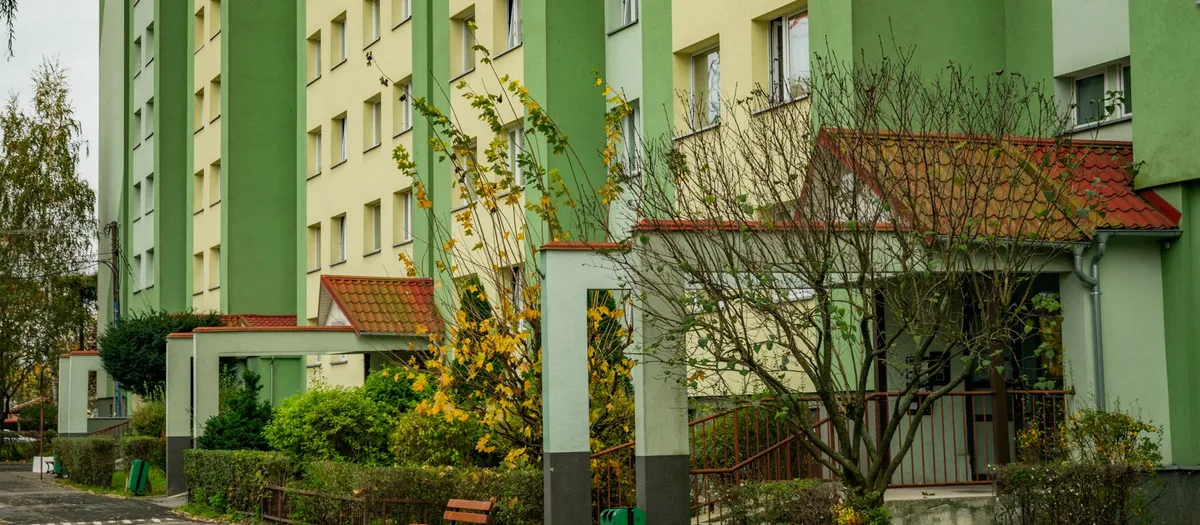 Sanierte Wohnblöcke in Grün und Creme mit überdachten Hauseingängen, davor Hecken, eine Bank und herbstliche Sträucher