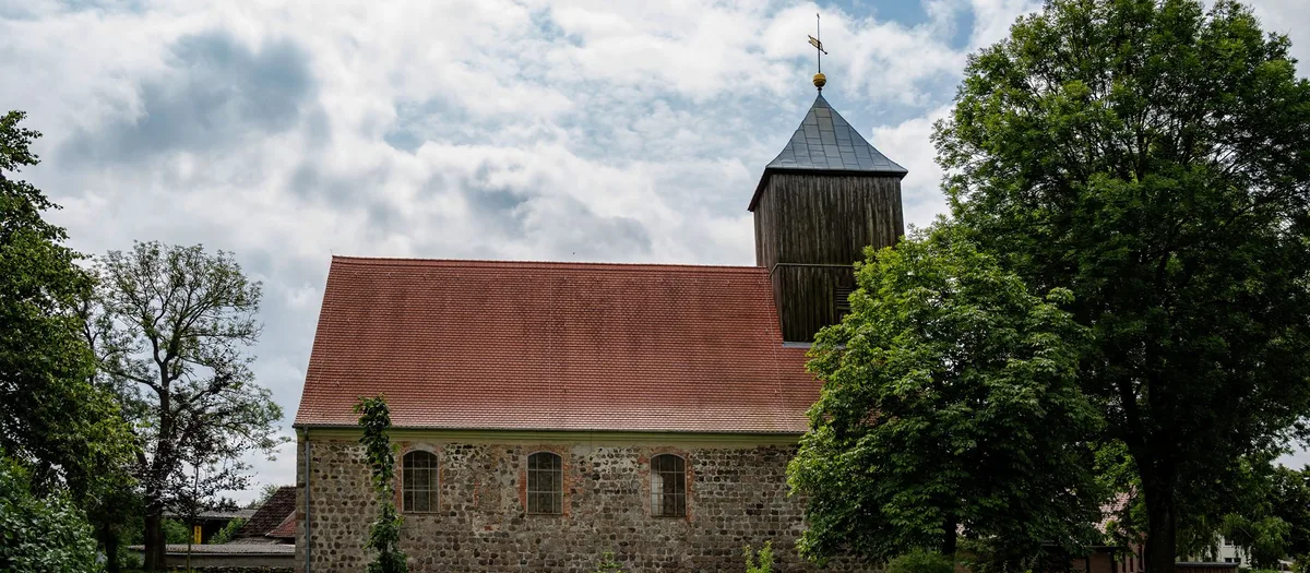 Dorfkirche aus Feldstein mit rotem Ziegeldach und hölzernem Glockenturm, umgeben von alten Laubbäumen