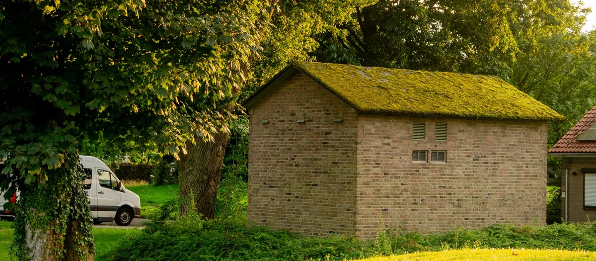 Gemauerter Schuppen mit bemoostem Dach in einem großen Garten, daneben Rasen und alte Laubbäume